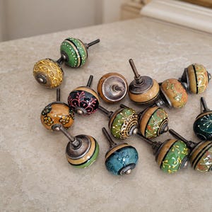 May include: A collection of twelve round cabinet knobs featuring various hand-painted designs in shades of green, yellow, blue, and brown. Each knob has a metallic silver-toned base and a threaded metal bolt extending from the back for installation.