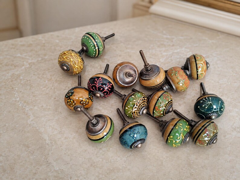 May include: A collection of twelve round cabinet knobs featuring various hand-painted designs in shades of green, yellow, blue, and brown. Each knob has a metallic silver-toned base and a threaded metal bolt extending from the back for installation.