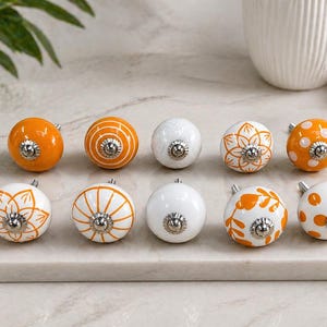May include: A collection of ten decorative cabinet knobs. The knobs are round with a variety of designs, including solid orange, white with orange stripes, and white with orange floral patterns. Each knob has a silver metal base.