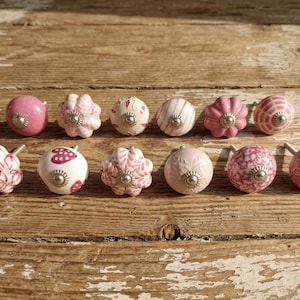 Puede incluir: Una colección de pomos de cerámica rosa y blanca con herrajes plateados. Los pomos presentan varios diseños florales y estampados, incluyendo rayas y lunares. Están dispuestos sobre una superficie de madera envejecida.
