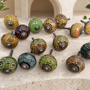 May include: A collection of thirteen round ceramic cabinet knobs featuring various hand-painted patterns in shades of green, yellow, orange, and black. Each knob has a central metallic hardware piece and a short metal bolt for installation.