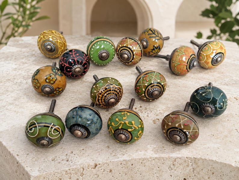 May include: A collection of thirteen round ceramic cabinet knobs featuring various hand-painted patterns in shades of green, yellow, orange, and black. Each knob has a central metallic hardware piece and a short metal bolt for installation.