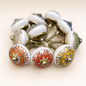 Pu&ograve; includere: Una collezione di dieci pomelli rotondi in ceramica per armadietti con vari motivi geometrici colorati in arancione, giallo, verde, blu e oro. Ogni pomello ha una base bianca, un accento centrale in metallo dorato e un bullone filettato.