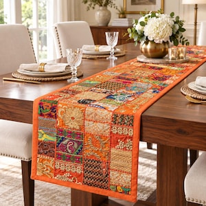 Op de afbeelding: Een lange rechthoekige tafelloper met een levendig patchworkontwerp van oranje, gouden en veelkleurige stoffen vierkanten. De loper is versierd met ingewikkeld borduurwerk, accenten van metaaldraad en kralenwerk, plat liggend op een houten tafel.
