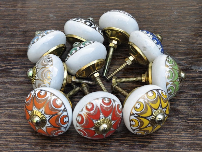 Pu&ograve; includere: Una collezione di dieci pomelli rotondi in ceramica per mobili con vari motivi geometrici colorati in arancione, giallo, verde e blu. Ogni pomello ha una base bianca, un raccordo in metallo color ottone e un bullone filettato per il montaggio.