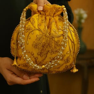 Puede incluir: Un bolso amarillo con bordados florales dorados y una correa de cuentas. El bolso tiene forma redonda, borlas doradas y adornos de cuentas perladas. El bolso es sostenido por una persona.