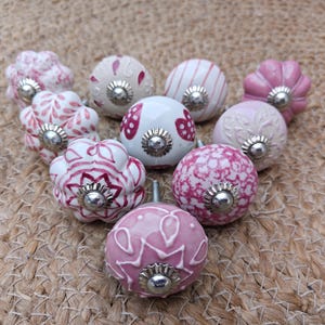 Puede incluir: Una colección de pomos de cerámica decorativos en varios tonos de rosa y blanco. Los pomos presentan diseños florales y estampados, con herrajes de metal plateado. Los pomos están dispuestos sobre una superficie tejida y texturizada.