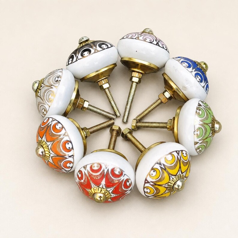 Pu&ograve; includere: Una collezione di otto pomelli rotondi in ceramica per mobili disposti in cerchio. Ogni pomello presenta una base bianca con motivi colorati distinti in arancione, giallo, verde, blu, rosso e oro metallizzato, con un bullone in ottone.