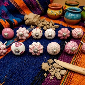 Könnte beinhalten: Eine Sammlung dekorativer Keramik-Schrankknöpfe in verschiedenen rosa- und weißen Designs, mit silberfarbener Hardware. Die Knöpfe sind auf einem farbenfrohen, gemusterten Textil angeordnet, mit kleinen Tonstücken und Miniatur-Töpferwaren im Hintergrund.