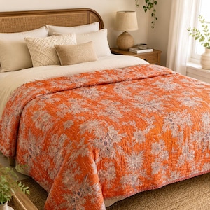 Könnte beinhalten: Eine leuchtend orangefarbene Steppdecke mit einem detaillierten Blumenmuster in gedeckten Weiß- und Grautönen. Die Decke weist durchgehende vertikale Steppnähte auf, die dem Stoff eine strukturierte, gesteppte Optik auf der gesamten Bettfläche verleihen.