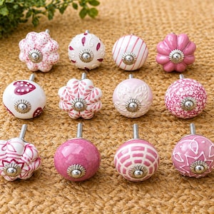 May include: A collection of decorative ceramic cabinet knobs in various shades of pink and white. The knobs feature floral and patterned designs, with silver-toned metal hardware. The knobs are arranged on a textured, tan surface.
