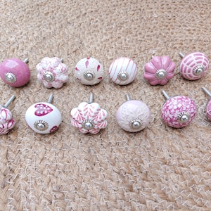 Könnte beinhalten: Eine Sammlung von zwölf dekorativen Schrankknöpfen in verschiedenen Rosa- und Weißtönen. Jeder Knopf weist ein einzigartiges Muster auf, darunter florale, gestreifte, herzförmige und geometrische Designs, jeweils mit einer silberfarbenen Schraube.