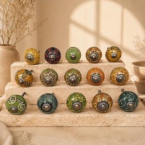 Op de afbeelding: Een collectie van vijftien ronde keramische kastknoppen gerangschikt op een stenen display met niveaus. Elke knop heeft een uniek handgeschilderd patroon in tinten groen, geel, oranje en zwart, geaccentueerd met metalen beslag in het midden.