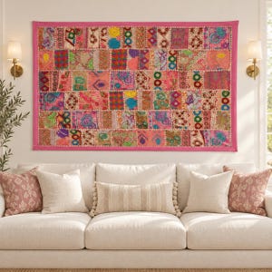 Peut inclure: Une tenture murale rectangulaire présentant un motif patchwork composé de divers carrés de tissu brodés dans des tons de rose, orange, vert et bleu, finie avec une bordure rose unie et des coutures décoratives sur toute la surface.