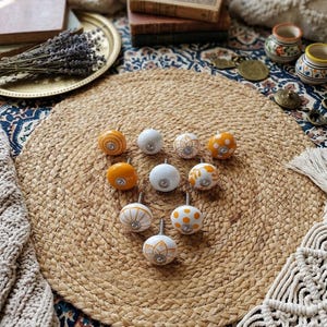 Pode incluir: Um conjunto de puxadores decorativos para armários em tons de laranja e branco, com desenhos florais e estampados. Os puxadores são exibidos sobre um tapete redondo de juta tecida, para decoração de casa.