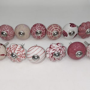 Peut inclure: Un ensemble de douze boutons de placard décoratifs disposés sur deux rangées. Chaque bouton présente un motif unique dans des tons de rose, blanc et crème, incluant des designs floraux, géométriques et des cœurs. Chaque pièce a une vis centrale.