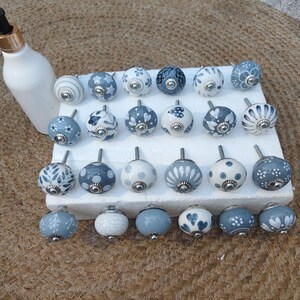 Op de afbeelding: Een verzameling decoratieve keramische kastknoppen in verschillende ontwerpen en kleuren, waaronder wit, blauw en grijs. De knoppen hebben bloemen-, stippen- en patroondetails. Een witte fles met een zwarte en gouden dop is ook aanwezig.