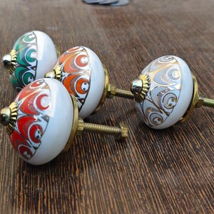 Pu&ograve; includere: Tre pomelli rotondi in ceramica per mobili con base bianca e accenti in oro metallizzato. Ogni pomello presenta un distinto motivo colorato in verde, arancione o rosso, con una base in metallo color ottone e un perno filettato sul retro.