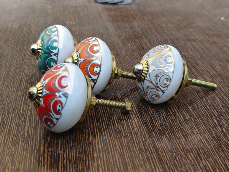 Pu&ograve; includere: Tre pomelli rotondi in ceramica per mobili con base bianca e accenti in oro metallizzato. Ogni pomello presenta un distinto motivo colorato in verde, arancione o rosso, con una base in metallo color ottone e un perno filettato sul retro.