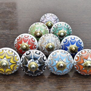 Pu&ograve; includere: Una collezione di dieci pomelli rotondi in ceramica per mobili disposti a triangolo. Ogni pomello presenta un motivo floreale multicolore unico con intricati dettagli a linee dorate e un elemento centrale in metallo dorato.