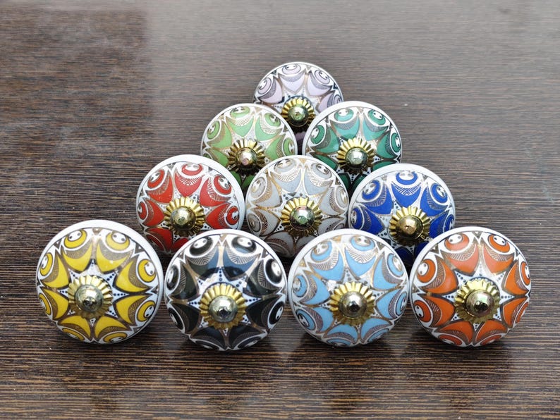 Pu&ograve; includere: Una collezione di dieci pomelli rotondi in ceramica per mobili disposti a triangolo. Ogni pomello presenta un motivo floreale multicolore unico con intricati dettagli a linee dorate e un elemento centrale in metallo dorato.