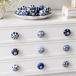 May include: A white chest of drawers with blue and white ceramic knobs. The knobs display floral and geometric patterns. A white ceramic bowl filled with matching knobs sits on top, along with books and a vase of flowers.