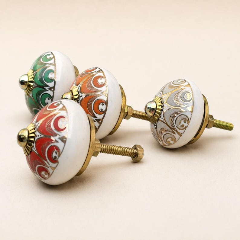 Pu&ograve; includere: Tre pomelli rotondi in ceramica per mobili con base bianca e motivi complessi ispirati alle piume di pavone in verde, arancione e grigio, accentuati da dettagli dorati e ferramenta in metallo color ottone con perno filettato e dado.