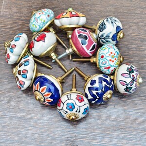 Pu&ograve; includere: Una collezione di pomelli decorativi in ceramica con ferramenta in ottone. I pomelli presentano vari disegni dipinti a mano in blu, rosso, bianco e altri colori. I pomelli sono disposti su una superficie di legno.