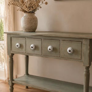 May include: A sage green wooden console table with four drawers, each featuring a unique white ceramic knob with a dark centre. The table has a lower shelf and turned legs. A beige ceramic vase filled with dried flowers sits on the top surface.