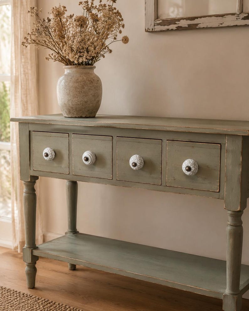 May include: A sage green wooden console table with four drawers, each featuring a unique white ceramic knob with a dark centre. The table has a lower shelf and turned legs. A beige ceramic vase filled with dried flowers sits on the top surface.