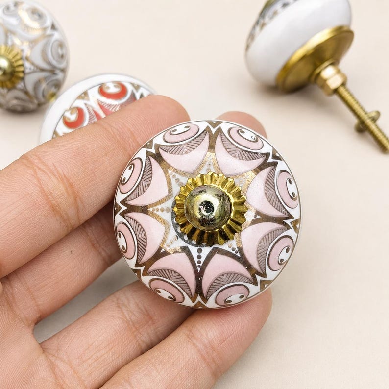 Pu&ograve; includere: Un pomello rotondo in ceramica per mobili con un motivo simmetrico di petali rosa tenue e bianchi con accenti metallici dorati. Il centro del pomello presenta un tappo metallico dorato smerlato e un bullone filettato sul retro.