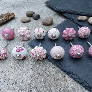 Könnte beinhalten: Eine Sammlung dekorativer Keramik-Schrankknöpfe in verschiedenen Rosa- und Weißtönen. Die Knöpfe weisen verschiedene Muster auf, darunter Blumen-, Streifen- und Punktmuster, mit silberfarbener Metallbeschlägen. Die Knöpfe sind auf einer strukturierten Oberfläche angeordnet.