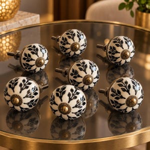 May include: A collection of seven round ceramic cabinet knobs featuring a white base with a black leaf pattern and a central antique brass-toned metal accent. Each knob includes a threaded metal bolt and a nut for secure installation on furniture.