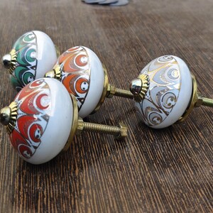 Pu&ograve; includere: Tre pomelli rotondi in ceramica per mobili con base bianca e accenti in oro metallizzato. Ogni pomello presenta un motivo colorato distinto in verde, arancione o rosso, con una base in metallo color ottone e una vite filettata per il montaggio.