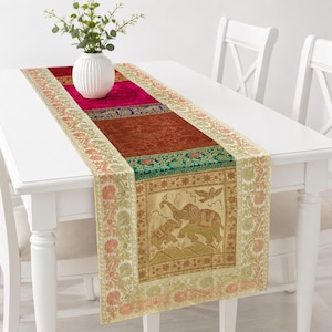 May include: A rectangular table runner featuring a patchwork design of vibrant magenta, deep rust, and cream panels. The fabric displays intricate woven patterns, including a central gold-toned section depicting two elephants and a bird.