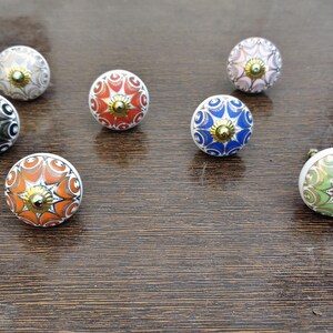 Pu&ograve; includere: Una collezione di sette pomelli rotondi in ceramica per mobili con intricati motivi floreali e geometrici multicolori nelle tonalit&agrave; arancione, blu, verde, nero e bianco, ciascuno sormontato da un piccolo perno centrale in metallo dorato lucido.