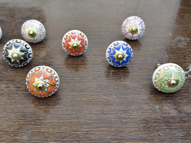 Pu&ograve; includere: Una collezione di sette pomelli rotondi in ceramica per mobili con intricati motivi floreali e geometrici multicolori nelle tonalit&agrave; arancione, blu, verde, nero e bianco, ciascuno sormontato da un piccolo perno centrale in metallo dorato lucido.