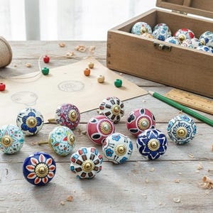 Può includere: Una collezione di pomelli decorativi in ceramica in vari colori e motivi, tra cui disegni floreali e geometrici. Alcuni pomelli sono esposti su una superficie di legno, altri all'interno di una scatola di legno aperta. Sono presenti anche una matita, un righello e dello spago.