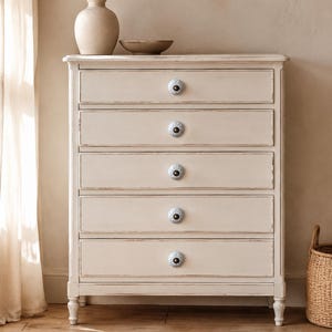 May include: A tall, off-white wooden chest of drawers featuring five horizontal drawers. Each drawer is equipped with a single, round, light blue ceramic knob with a detailed floral pattern and a dark centre, set against the distressed cream finish.