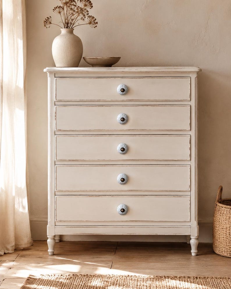 May include: A tall, off-white wooden chest of drawers featuring five horizontal drawers. Each drawer is equipped with a single, round, light blue ceramic knob with a detailed floral pattern and a dark centre, set against the distressed cream finish.