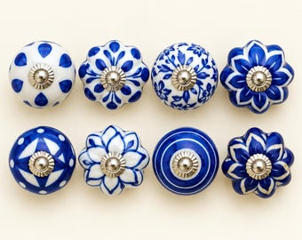 Może przedstawiać: Zestaw ośmiu okrągłych ceramicznych gałek do szafek ułożonych w dwóch rzędach. Każda gałka posiada unikalny niebiesko-biały wzór, w tym motywy kwiatowe, geometryczne i pasy. Każda gałka ma centralnie umieszczony srebrny metalowy element montażowy.