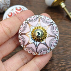 Pu&ograve; includere: Un pomello rotondo in ceramica per mobili con base bianca e un motivo simmetrico di petali rosa tenue, linee grigio scuro e accenti dorati. Il centro presenta un tappo in metallo dorato smerlato con un piccolo rivetto metallico riflettente.