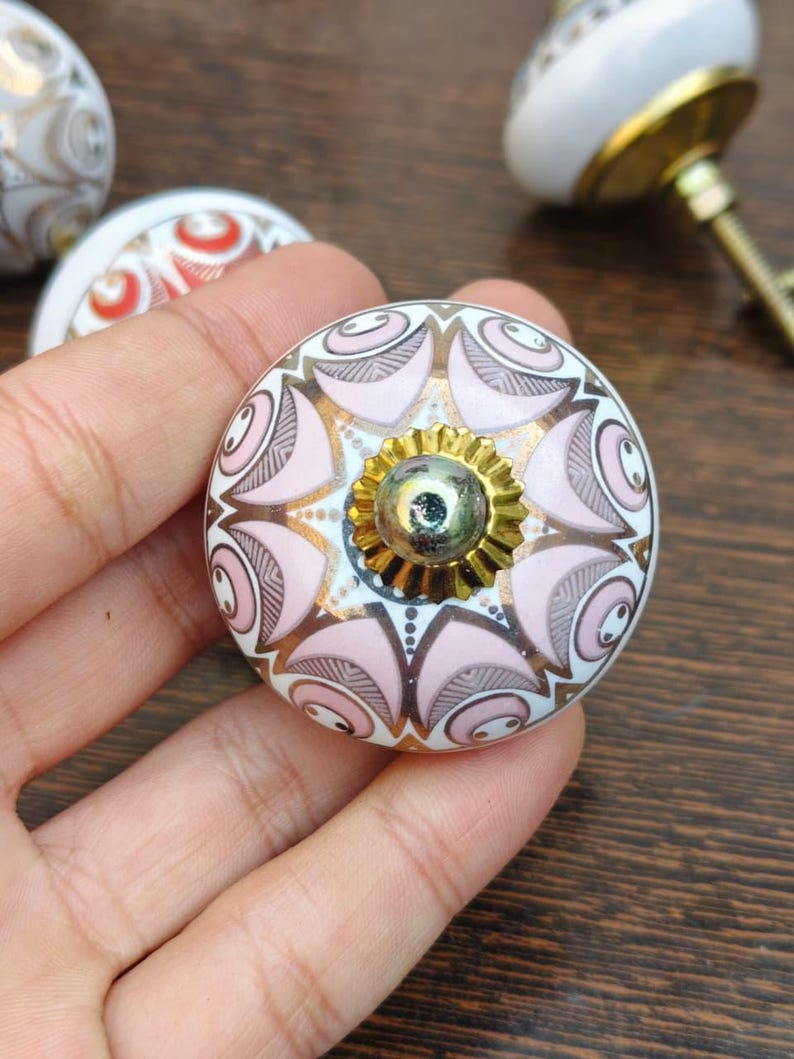 Pu&ograve; includere: Un pomello rotondo in ceramica per mobili con base bianca e un motivo simmetrico di petali rosa tenue, linee grigio scuro e accenti dorati. Il centro presenta un tappo in metallo dorato smerlato con un piccolo rivetto metallico riflettente.