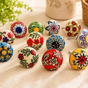 May include: A collection of eleven round ceramic knobs featuring various hand-painted floral and geometric patterns in vibrant colors including red, blue, yellow, green, and white. Each knob has a metal screw attachment extending from the back.