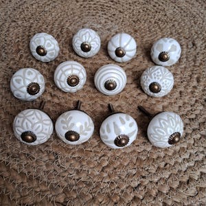 May include: A collection of twelve round ceramic cabinet knobs featuring a white base with light beige patterns and a central antique bronze-coloured metal hardware piece. Each knob displays a unique geometric or floral design on its surface.