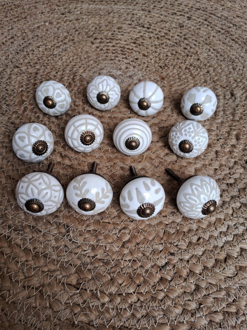 May include: A collection of twelve round ceramic cabinet knobs featuring a white base with light beige patterns and a central antique bronze-coloured metal hardware piece. Each knob displays a unique geometric or floral design on its surface.