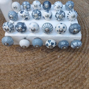 Op de afbeelding: Een verzameling decoratieve keramische kastknoppen in verschillende ontwerpen. De knoppen zijn voornamelijk wit en blauw, met bloemen- en patroondetails. Elke knop heeft een zilverkleurige metalen basis en is gerangschikt op een wit oppervlak.