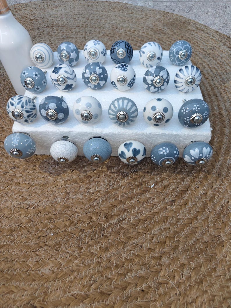 Op de afbeelding: Een verzameling decoratieve keramische kastknoppen in verschillende ontwerpen. De knoppen zijn voornamelijk wit en blauw, met bloemen- en patroondetails. Elke knop heeft een zilverkleurige metalen basis en is gerangschikt op een wit oppervlak.