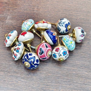 Pu&ograve; includere: Una collezione di pomelli decorativi in ceramica con finiture in ottone. I pomelli presentano vari motivi floreali e geometrici colorati in rosso, blu, bianco e giallo. I pomelli sono disposti su una superficie di legno.