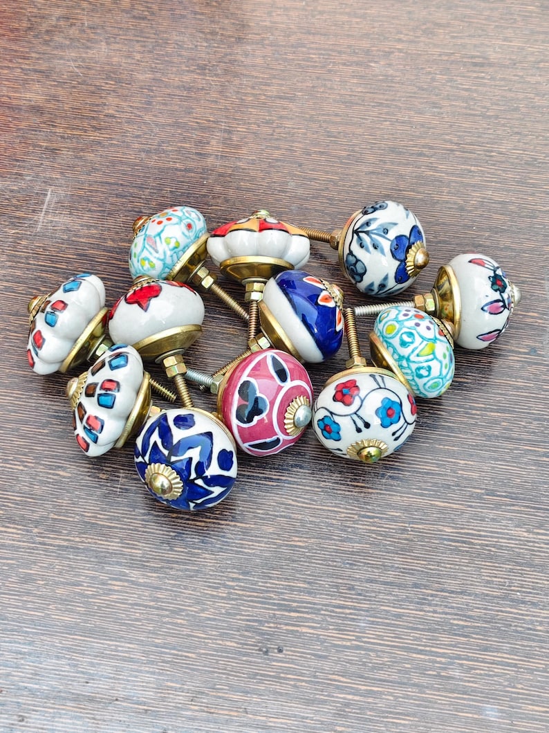 Pu&ograve; includere: Una collezione di pomelli decorativi in ceramica con finiture in ottone. I pomelli presentano vari motivi floreali e geometrici colorati in rosso, blu, bianco e giallo. I pomelli sono disposti su una superficie di legno.